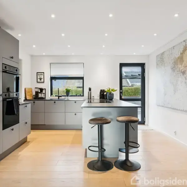 Et moderne køkken viser en central ø med to barstole. Et stort verdenskort hænger på væggen. Rummet er lyst med grå skabe og store vinduer.