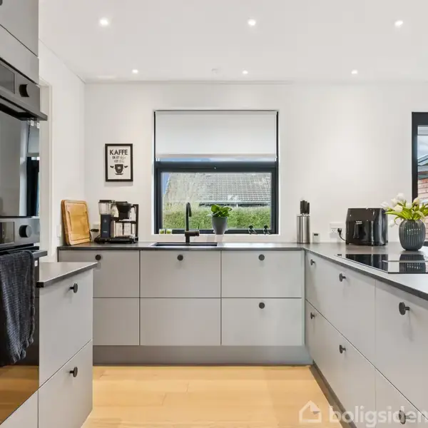 Køkken med grå skabe og sorte bordplader, kaffemaskine og planter på bordet. En bageplade hænger ved siden af ovnen. Vinduer viser udsigt til en have. Skilt: "KAFFE fordi...".