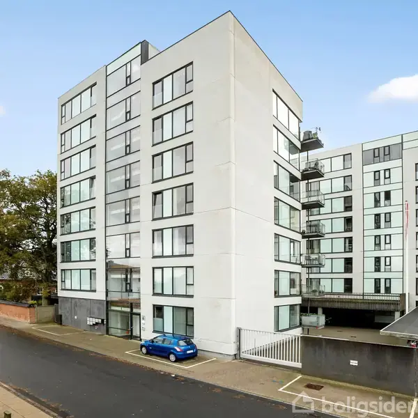 En moderne boligblok med flere etager står stille langs en gade. Ved siden af bygningen holder en blå bil parkeret på fortovet. Træer og andre bygninger ses i baggrunden.