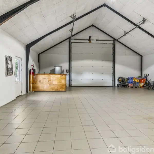 Garage med højt loft, biler parkeret langs væggen, Red Bull-barstol i forgrunden, trædisk til venstre, stor port i baggrunden, gulvet dækket med grå fliser.