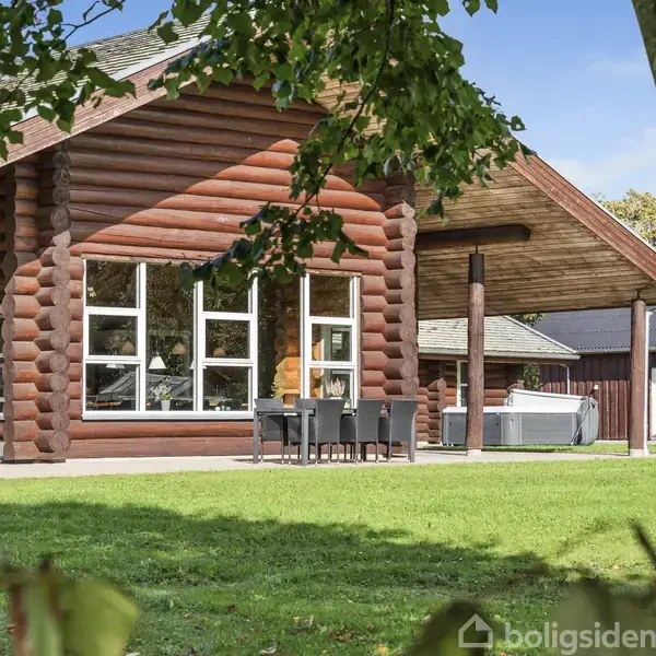 Træhus står stille i en grøn have, omgivet af græsplæne og træer. Ved siden af huset ses et udendørs spisebord med stole og en jacuzzi på terrassen.