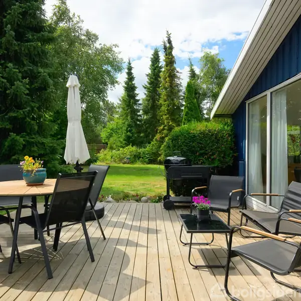 Terrassemøbler står arrangeret omkring et rundt bord med en parasol. Glidende skydedøre fører til et hus med blå ydervægge, omgivet af frodig have med høje træer.