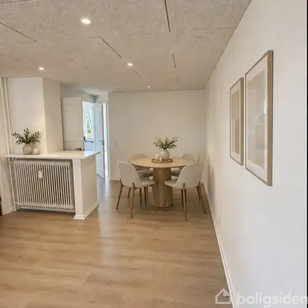 Et rundt spisebord står i hjørnet med fire stole omkring det. En vase med planter pryder bordet. Lys væg med to billeder og en åbent dør til venstre.