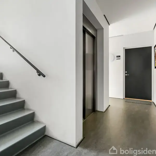 Trappeopgang med grå trin til venstre leder til en etage med elevator til højre. Dør ved siden af elevatoren. Vægplakater hænger til højre i moderne interiør.