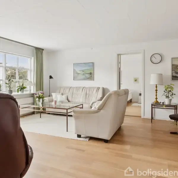 En stue med hvide sofaer omkring et glasbord. Store vinduer lader naturligt lys ind, mens planter og kunst pryder væggene. En åben dør fører til et andet rum.