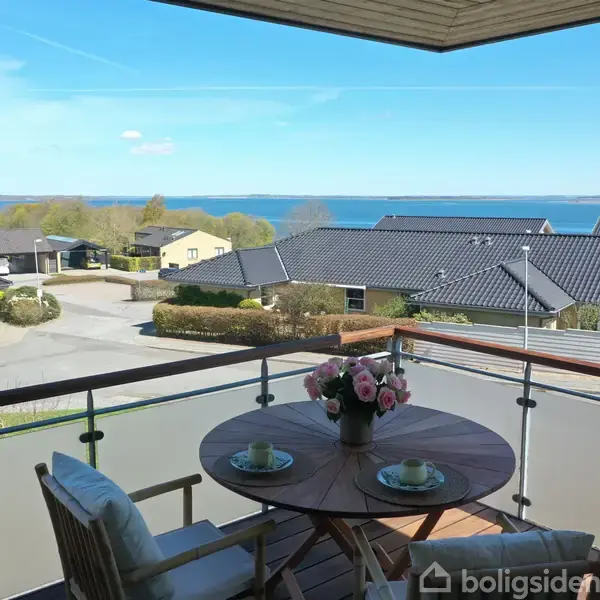 Runde bord med blomsterdekoration står på altan, to stole omkring. Udsigt over boligområde med huse og træer, hav i baggrunden under blå himmel.