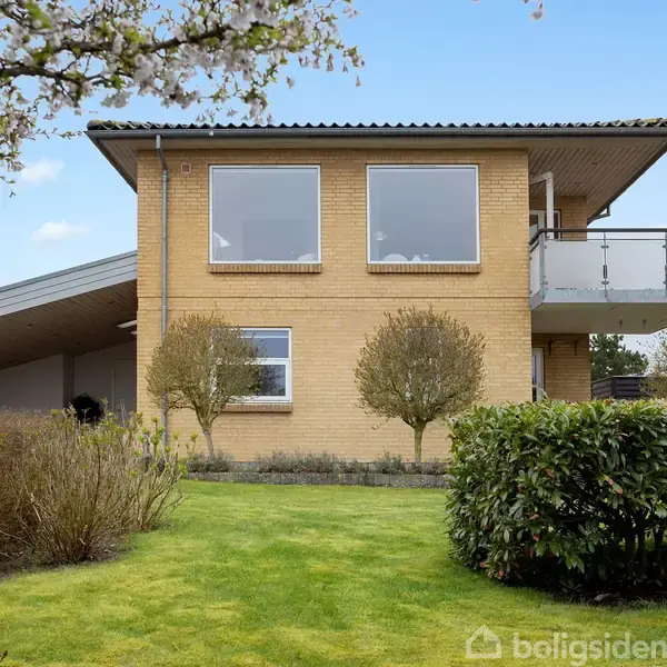 Et moderne tofamiliers hus i gule mursten står omgivet af en grøn have. To store vinduer og en balkon ses på førstesalen. Blomstrende træer rammer scenen.