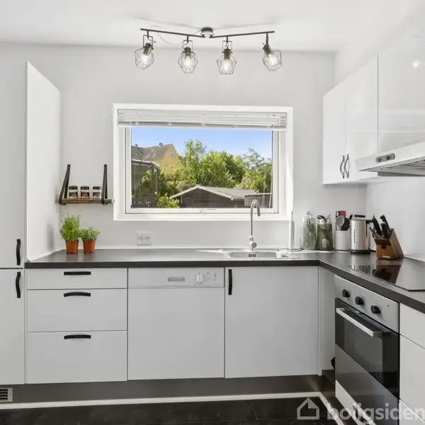 Et moderne køkken med hvide skabe og sorte bordplader. Rummet har en stor vindue, der slipper naturligt lys ind. Køkkenredskaber og krydderier står på bordpladen. Ingen tekst.