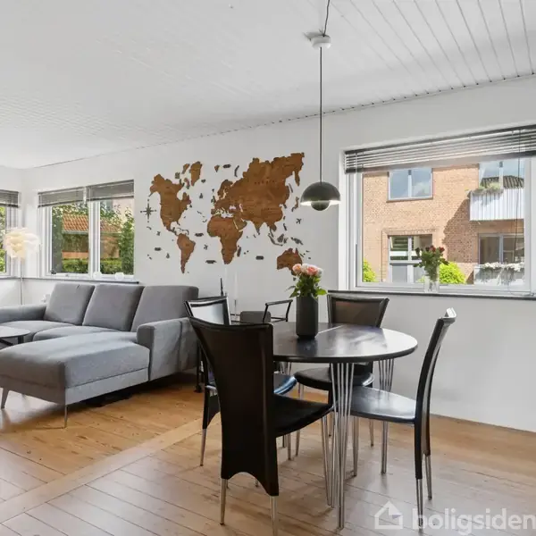 Gråt hjørnesofaarrangement står i stuen, foran en væg med verdenskort. Rundt sort spisebord med stole i centrum, omgivelser med trægulve og vinduer med udsigt til have.