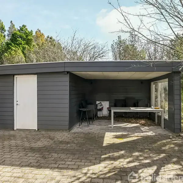 En grå bygning med fladt tag står på en flisebelagt terrasse. Bygningen har en hvid dør og store vinduer. Omgivelserne inkluderer træer og buske i en have.