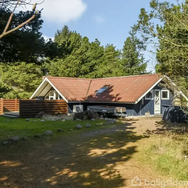 Et sommerhus står mellem træer og buske. Huset har rødbrunt tag og en lille terrasse. Omgivet af grønne områder på en solrig dag.