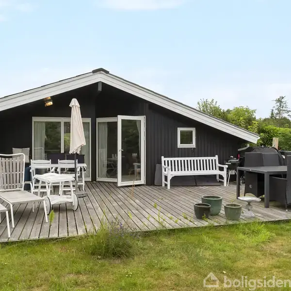 Sort træhus med åbne franske døre står på en træterrasse. Terrassen har hvide havemøbler, et hegn og planter i krukker. Græsplæne og buske omgiver huset.
