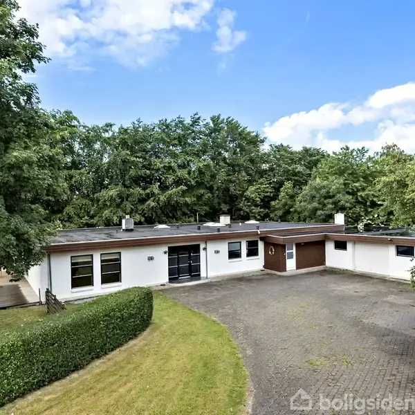 Et enfamilieshus med fladt tag står midt i en stor, brolagt gårdsplads omgivet af tæt træbevoksning og grønne plæner under en blå himmel med få skyer.