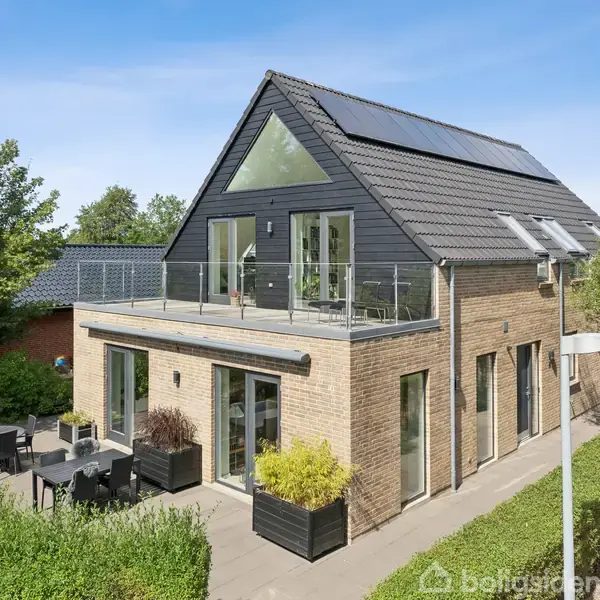 Hus med solpaneler på taget, omgivet af have og terrasse med havemøbler; lyse mursten og moderne design i et landområde med træer.