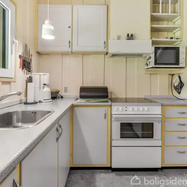 Et gulmalet træhus i skoven med en hvid bænk og vandkande foran. Omkring huset er der frodig vegetation og en stenlagt terrasse.