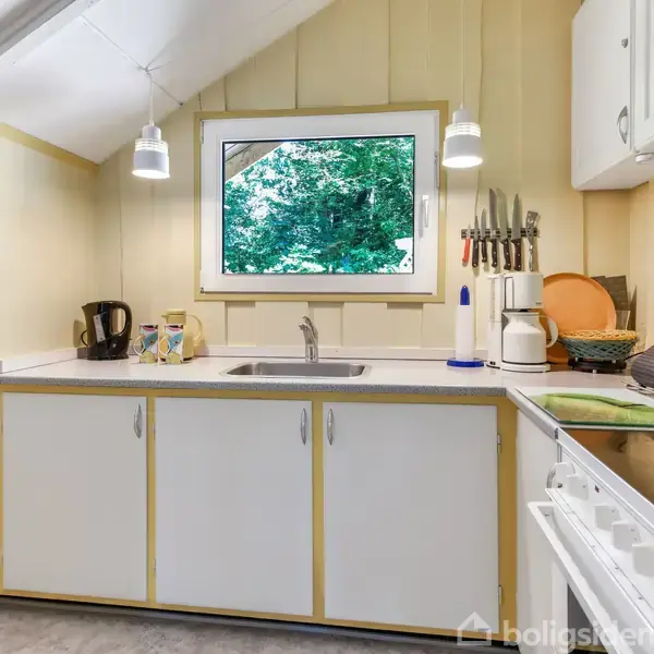 Et gult træhus med hvid dør og vindue står i en skovklædt have. En bænk og en vandkande er placeret på en brostensbelagt terrasse omgivet af frodig vegetation.