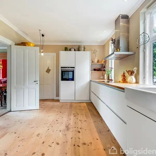 Køkken med hvide skabe og indbyggede apparater, stående på lyst trægulv, med udsigt til en grøn have gennem store vinduer, og adgang til en spisestue via åben dør.