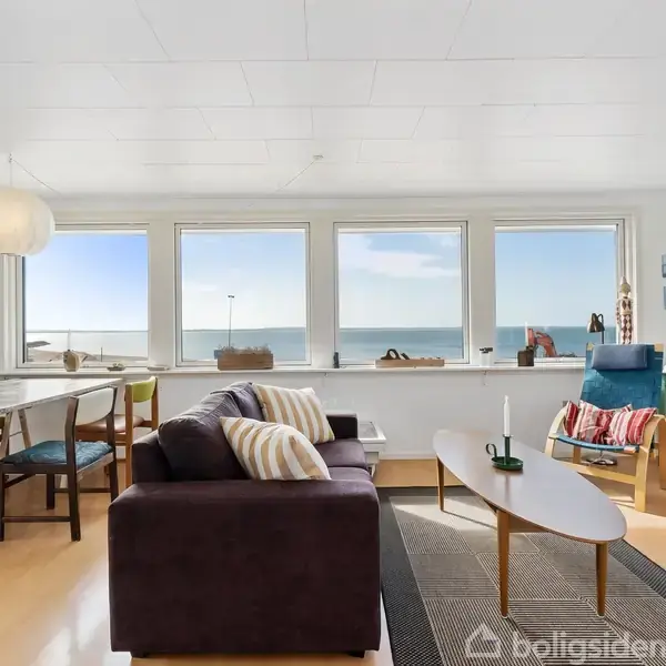 En stue indeholder en mørk sofa med pyntepuder, vendt mod et sofabord. Store vinduer giver udsigt til havet. I hjørnet er spisebord og stole, skaber en lys atmosfære.