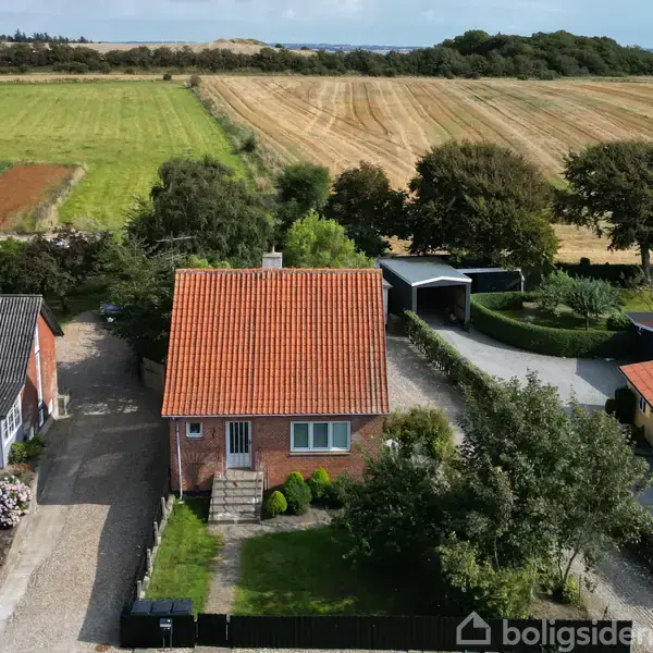 Et rødt murstenshus med rødt tegltag står i en landsby. Omgivet af grønne marker og træer. En indkørsel og en carport er synlige ved siden af huset.