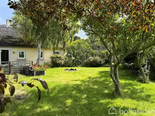 Et gulmalet hus er beliggende i en frodig have med grøn græsplæne og træer. En terrasse med blomsterkasser ses på venstre side, omgivet af tæt bevoksning og buske.