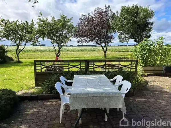 Et bord dækket med en dug står udenfor med fire hvide plaststole omkring det på en stenbelagt terrasse. I baggrunden er en grøn have med træer og marker.