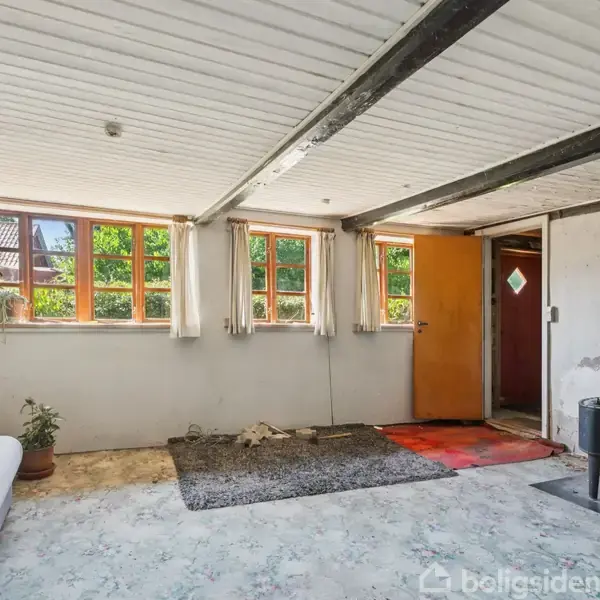 Et rum med en sofa og planter står ved siden af to vinduer med gardiner, der lukker lys ind. En åben dør fører til et andet rum, og en ældre brændeovn står til højre. Gulvet er dækket af forskellige tæpper.