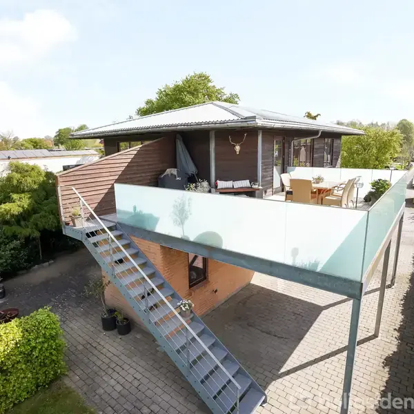 Et moderne hus med en stor terrasse, hvorpå der er placeret stole og bord. En metaltrappe fører op til terrassen. Grønne hække og træer omkranser ejendommen.
