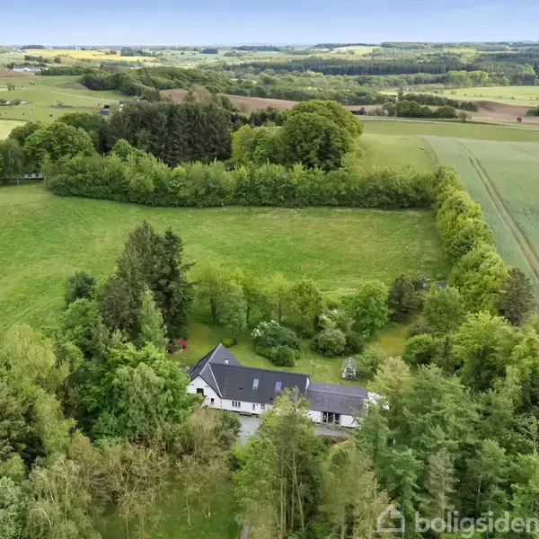 Et hus ligger omgivet af frodige grønne marker og tæt skov. Det er en landlig scene med vidtrækkende udsigt over det omkringliggende landskab.
