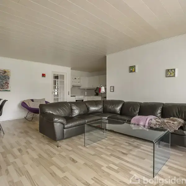 Sort lædersofa står i en stue med trægulv, forbundet til en glasbord. I baggrunden ses et spisebord med stole og et hvidt køkken. Vægpynt og belysning dekorerer rummet.