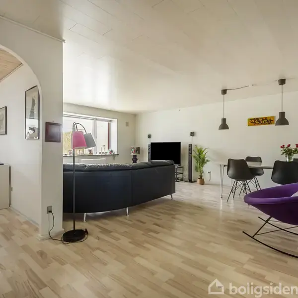 Stue med sort hjørnesofa og lilla lænestol, vendt mod et tv på væggen. Spisebord med stole ved siden af. Lyse trægulve og malerier pryder væggene.