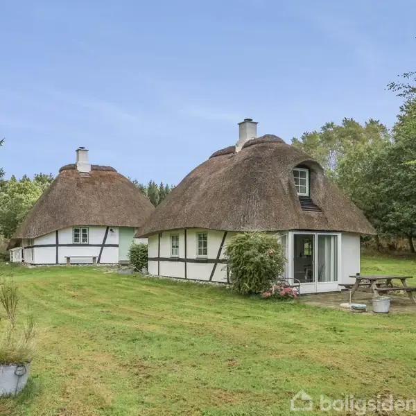 To stråtækte huse står i en grøn have med træer omkring. Foran det ene hus er der et picnicbord. Himlen er blå og området ser fredeligt ud.