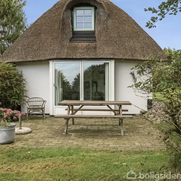 Stråtækt hus med en stor glasdør. Foran står et træbord med bænke. Huset er omgivet af en grøn have med buske og træer.