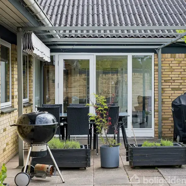 Grill står på en flisebelagt terrasse ved et rækkehus. Omkring den er flere plantekasser med grønne planter. I baggrunden ses havesæt og store glasdøre til huset.