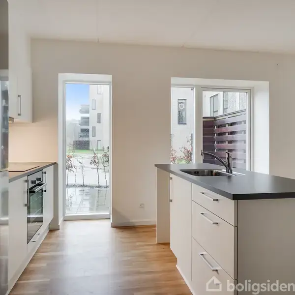 Køkken med hvidt skab og sort bordplade står tomt. Ovn og køleskab er indbygget til venstre. Store vinduer bagtil giver lys i rummet med trælaminatgulv.