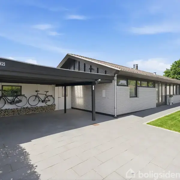 En moderne villa med hvid facade har en carport med to cykler monteret på væggen. Huset er omgivet af en flisebelagt indkørsel og en velplejet græsplæne samt grønne buske. Nummeret 230 vises.