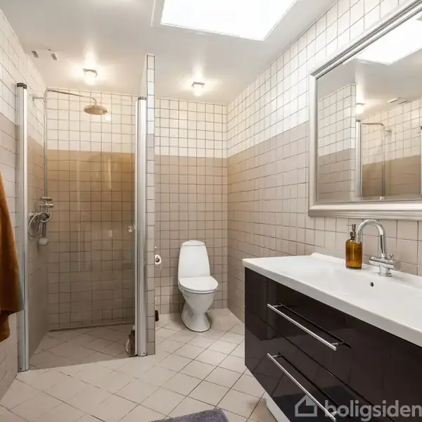 Badeværelse med brusekabine og toilet. Mørk skabsmodul med vask og spejl på højre side. Håndklæder hænger på væggen. Flisebelagt gulv og vægge.