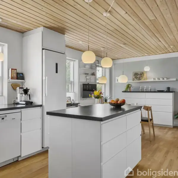 Et moderne køkken med hvide skabe og sort bordplade; loftet er beklædt med træ. Der hænger runde lamper, og planter er placeret på skabe. Gule blomster og frugter pynter på bordet.