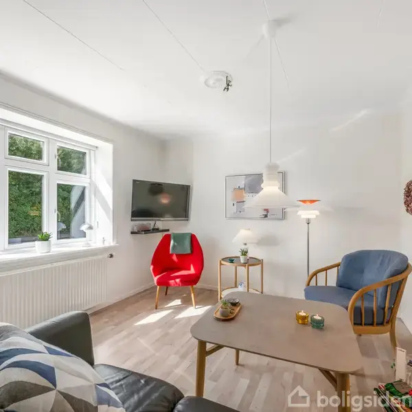 En lys stue indeholder en rød stol ved et vindue, en blå stol, et sofabord med pynt, og en fladskærm på væggen. Vinduerne viser grønne buske udenfor.