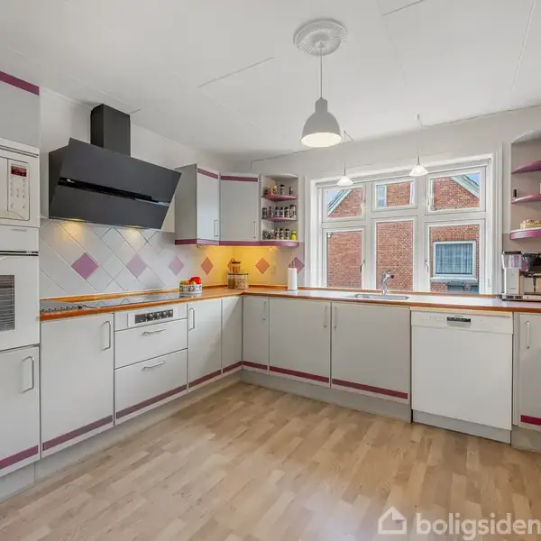 Et hvidt køkken med pink accenter har en indbygget ovn og mikrobølgeovn. En emhætte hænger over komfuret. Et vindue skaber et lyst rum med trægulve og hængende lamper.