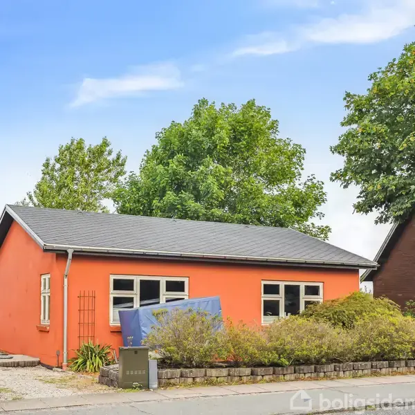En lille, orange hus bygning står stille omgivet af frodige træer og buske; en hæk grænser haven. I forgrunden ses en stille vej.