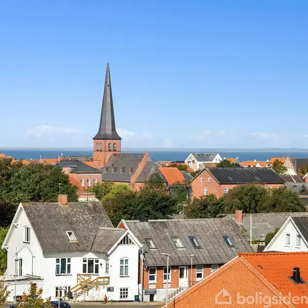 Kirketårn står centralt i en by med røde tage, omgivet af huse og træer. I baggrunden ses havet under en klar blå himmel.