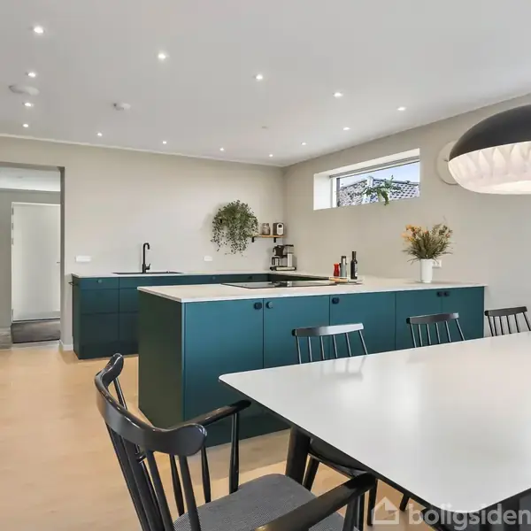 Et moderne køkken med mørkegrønne skabe og en hvid bordplade. Bord og stole dominerer spisestuen, mens en stor lampeskærm hænger over bordet. Rummet er lyst og minimalistisk indrettet.