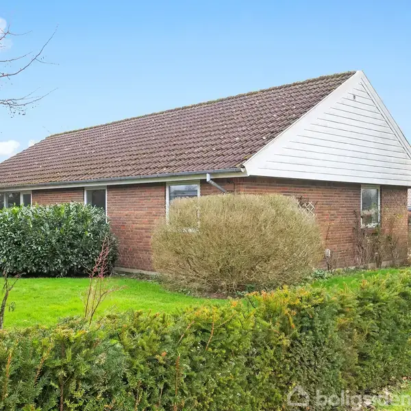 En rød murstenshus med tegltag er omgivet af en velplejet have med buske og græsplæne. Skydningen er i dagslys og viser en rolig forstadslignende beliggenhed.