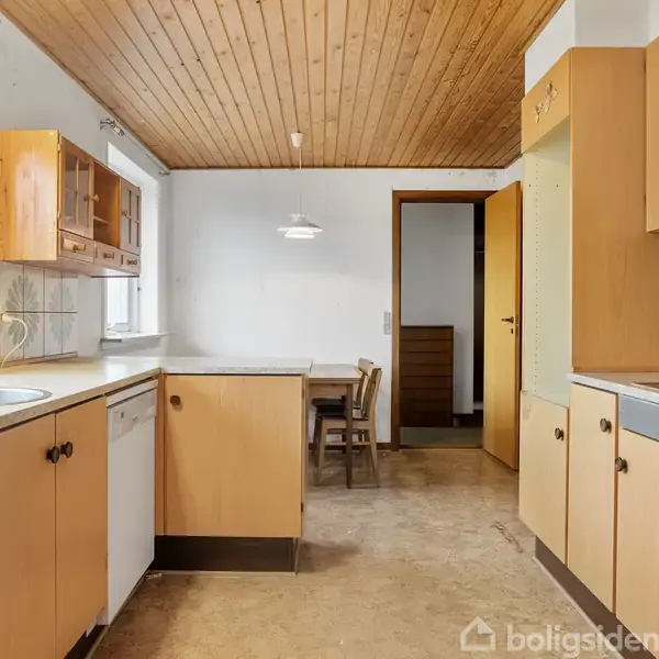 Køkken med træloft og lyse træskabe, hvor et spisebord med stole står i baggrunden. Fliser med blomsterdesign pynter væggene ved køkkenbordet. En åben dør leder til et andet rum.