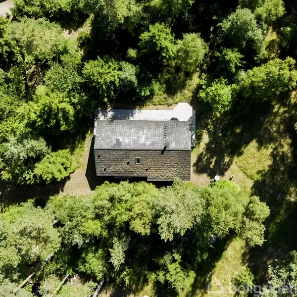 Et rektangulært hus ligger stille omgivet af frodig, tæt skov, der skaber en grøn og privat atmosfære. Huset har et mørkt tag og er omkranset af træer.