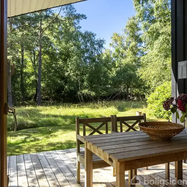 Træbord med blomsterkrukke og kurv står stille på terrasse. Udsigt over græsplæne og skov i dagslys, set gennem åben dør.