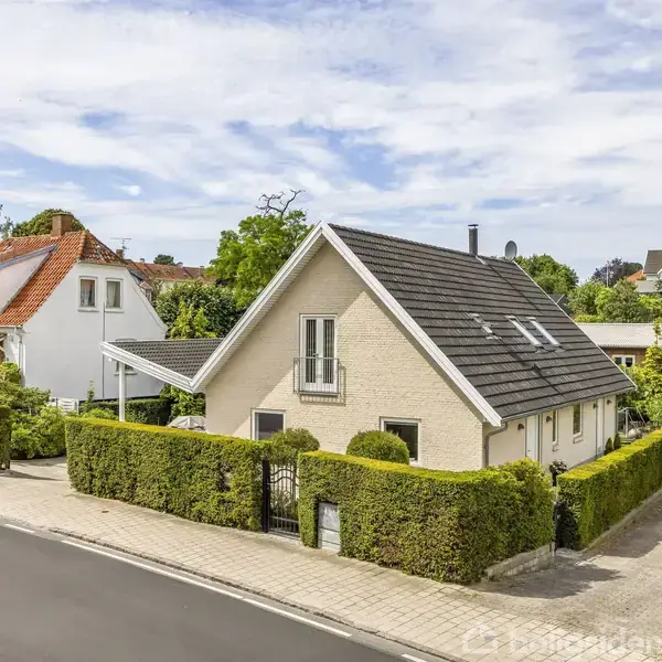 Et gult hus med gråt tag står på en hjørnegrund, omgivet af en grøn hæk. Det ligger langs en rolig gade med fortov og naboereder.