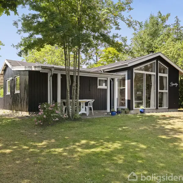 Sort træhus står på en solrig græsplæne omgivet af træer. Huset har store vinduer og en terrasse med et lille bord og stole. Navnet "Sysley" ses på facaden.
