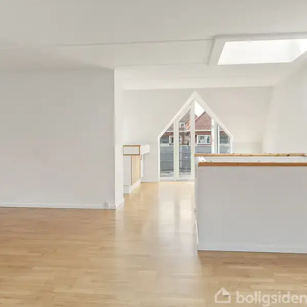 Rummelig, lys stue med parketgulv, åbent til entré med hylde. Store vinduer bagerst åbner ud mod en balkon, der giver udsigt over byen.