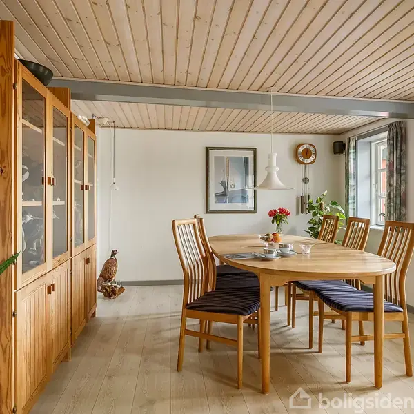 Et rundt træbord står med blomster på midten. Bagved ses træstole og en skænk langs væggen. Rummet har lyse trælofter, billeder på væggen og store vinduer.